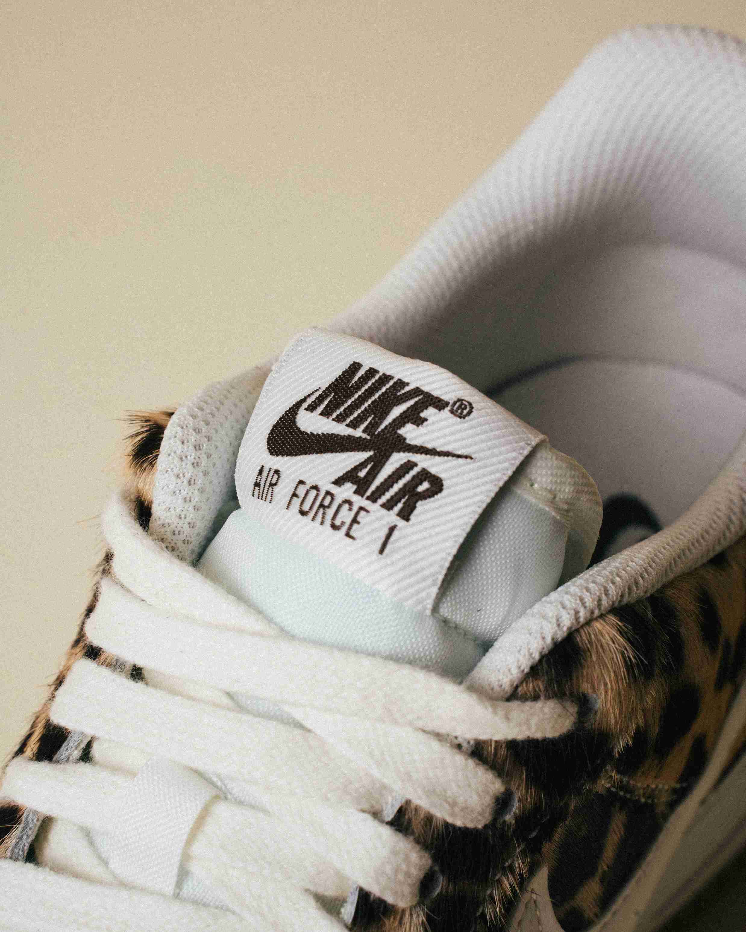 Paire de sneakers Nike Air Force 1 à motif léopard, placée à côté d’une boîte Nike grise sur un fond beige, vue artistique avec lumière douce.